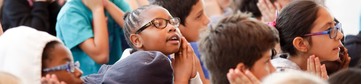 Banner photo of elementary students seated closely together, listening attentively; a student in the center wearing glasses clasps their hands near their face while looking forward.