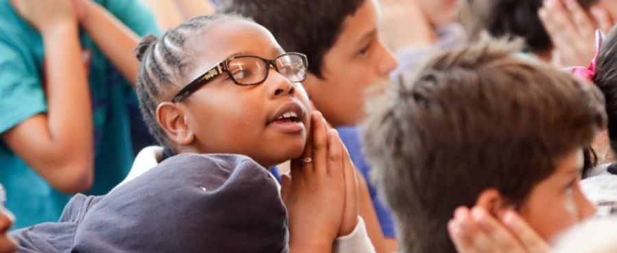 Banner photo of elementary students seated closely together, listening attentively; a student in the center wearing glasses clasps their hands near their face while looking forward.
